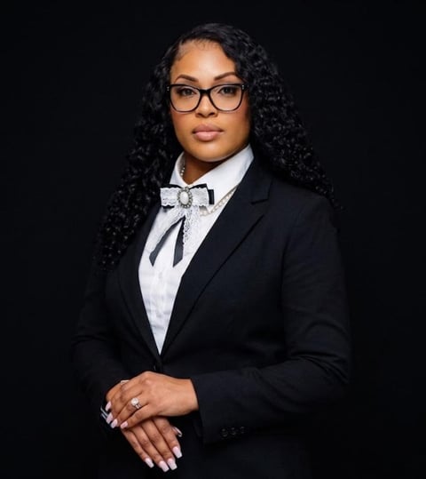 Professional headshot of woman wearing black blazer, white shirt with bow tie, and glasses against black background